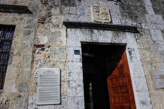 Entrada de la sede con escudo de armas, placa informativa y puerta de madera